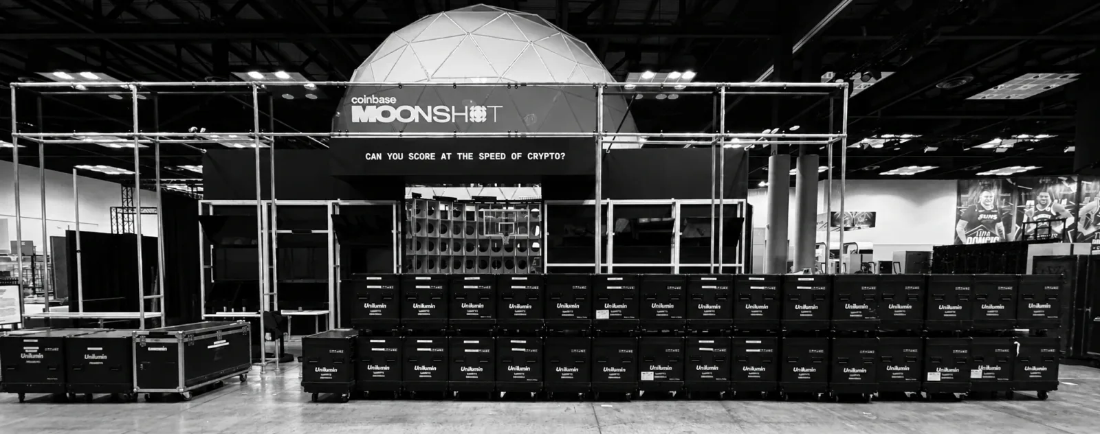 Equipment cases and staging framework beneath a geodesic dome at the Coinbase Moonshot installation, photographed in monochrome.