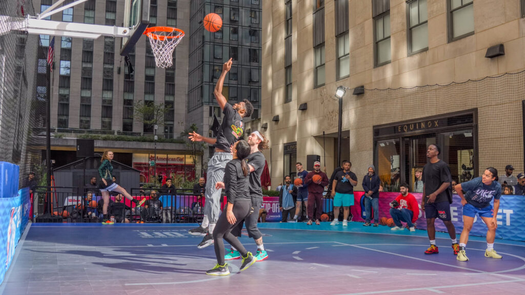 Basketball players reach for the hoop as spectators watch from beside the court.