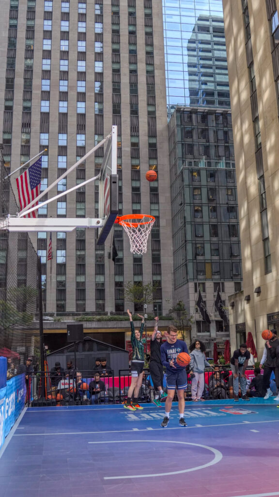 A basketball player approaches the hoop with Rockefeller Center buildings behind the court.